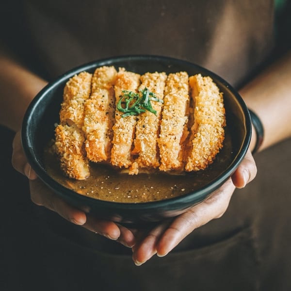 Hands holding a black bowl of golden-brown crispy chicken katsu with curry sauce and green garnish.